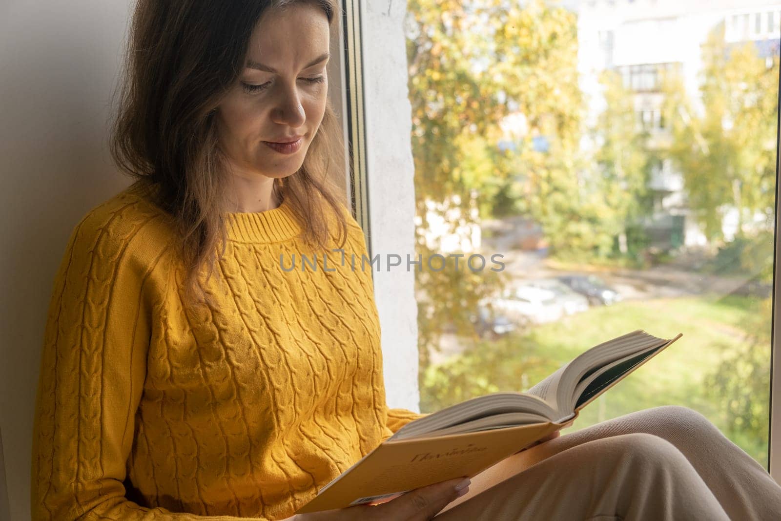 Young beautiful woman sitting by the window yellow knitted sweater read book, daily planner, notepad. Relax concept. Hold cappuccino glass of coffee with white foam. Text is out of focus
