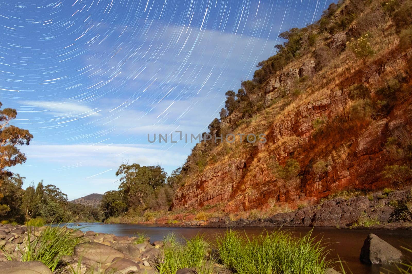 Stars trail across the night sky with a red rock gorge framing a running river in Australia