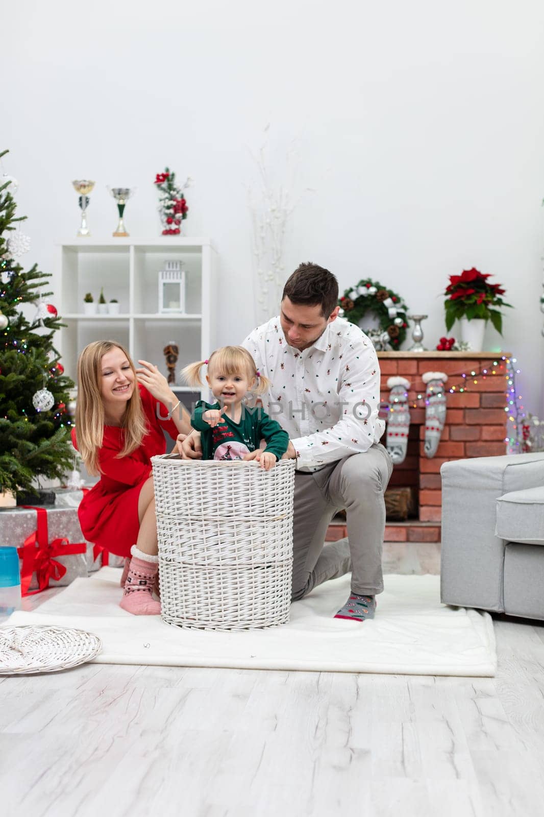Mom and dad are playing with their little daughter. The little girl has climbed into a large white wicker basket for fun. The family is smiling. In the background you can see a brick fireplace decorated with Christmas lights.