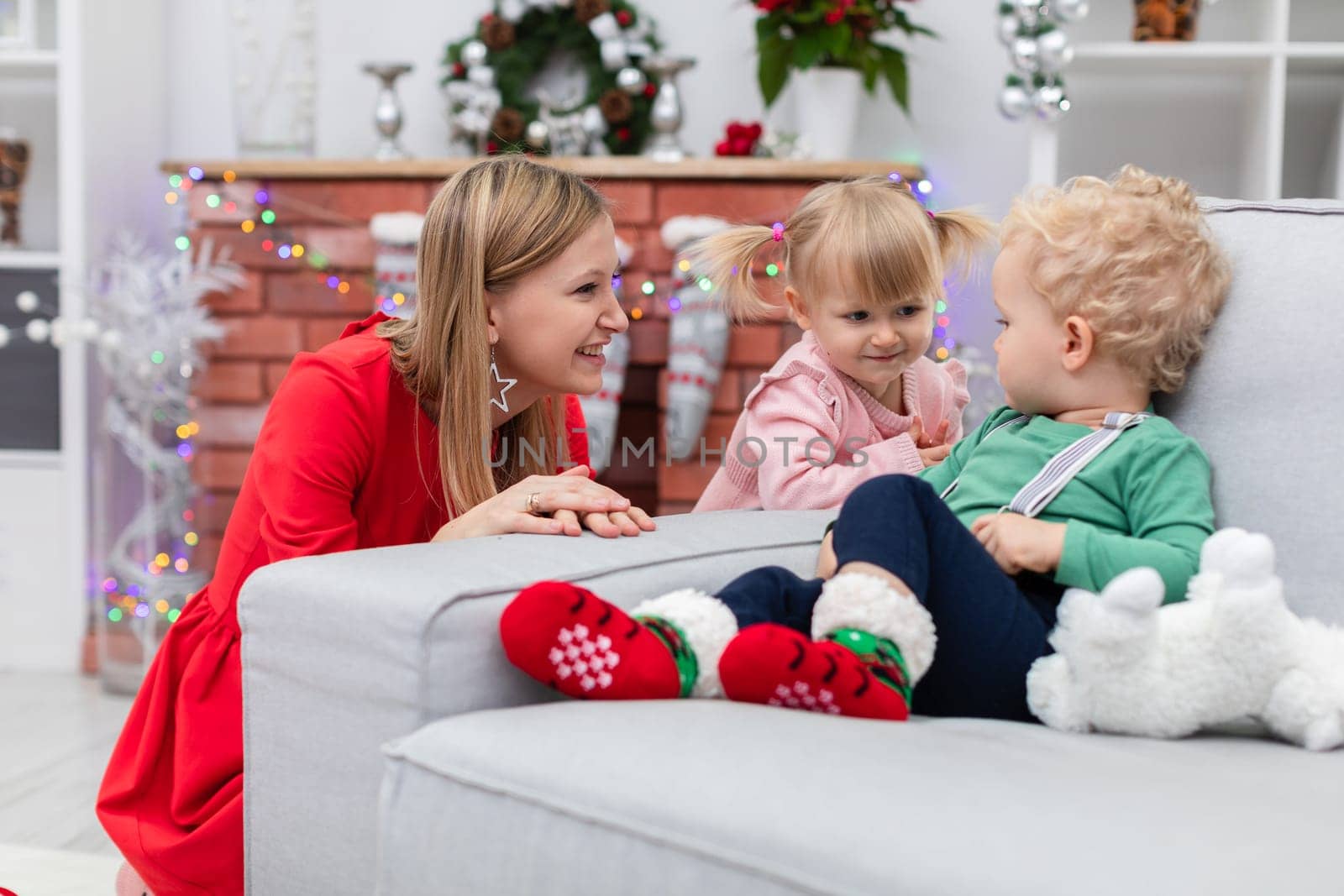 A little girl dressed in a pink dress stands by the edge of the couch. The girl looks at the boy, who is sitting on the couch. Next to the sibling is the mother. The woman crouches by the edge of the couch.