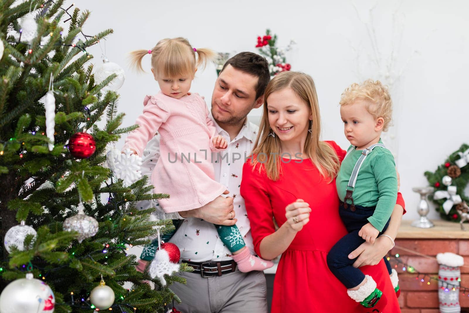 A little girl extends her hand toward the Christmas tree ornaments. A dad is holding his daughter in his arms. A woman in a red dress stands next to him. The woman holds a little boy in her arms.