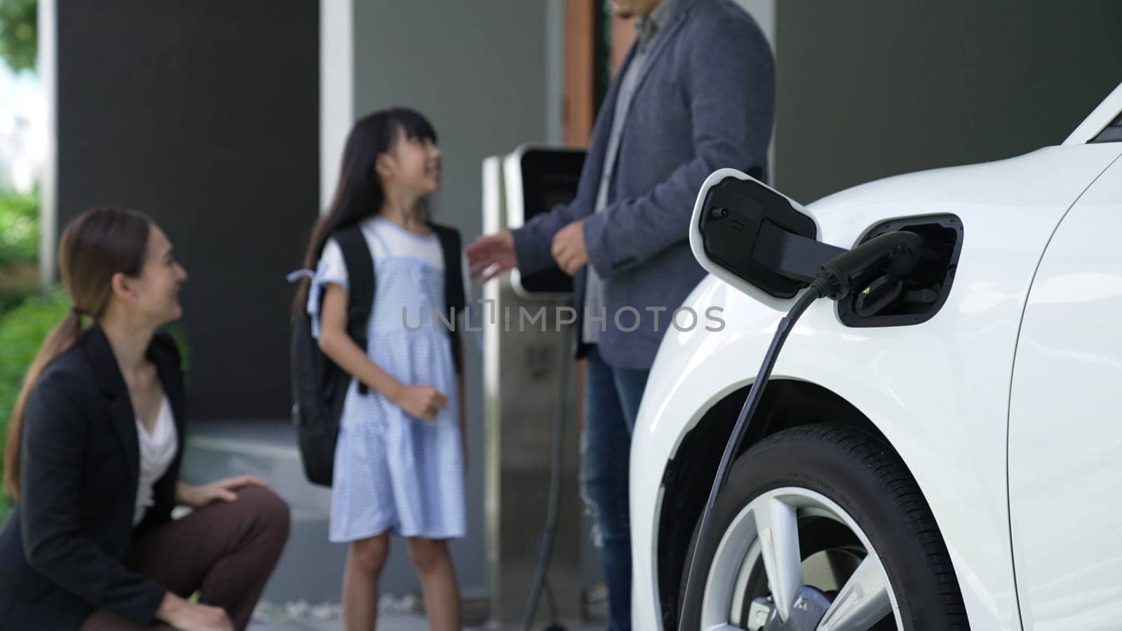 Progressive parents with electric vehicle and home charging station. Happy family with daughter giving each other high fives before leave for school. Alternative future transportation concept of EVs