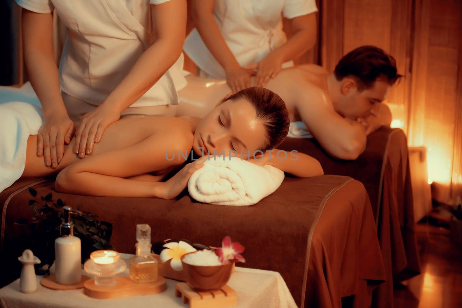 Caucasian couple customer enjoying relaxing anti-stress spa massage and pampering with beauty skin recreation leisure in warm candle lighting ambient salon spa at luxury resort or hotel. Quiescent
