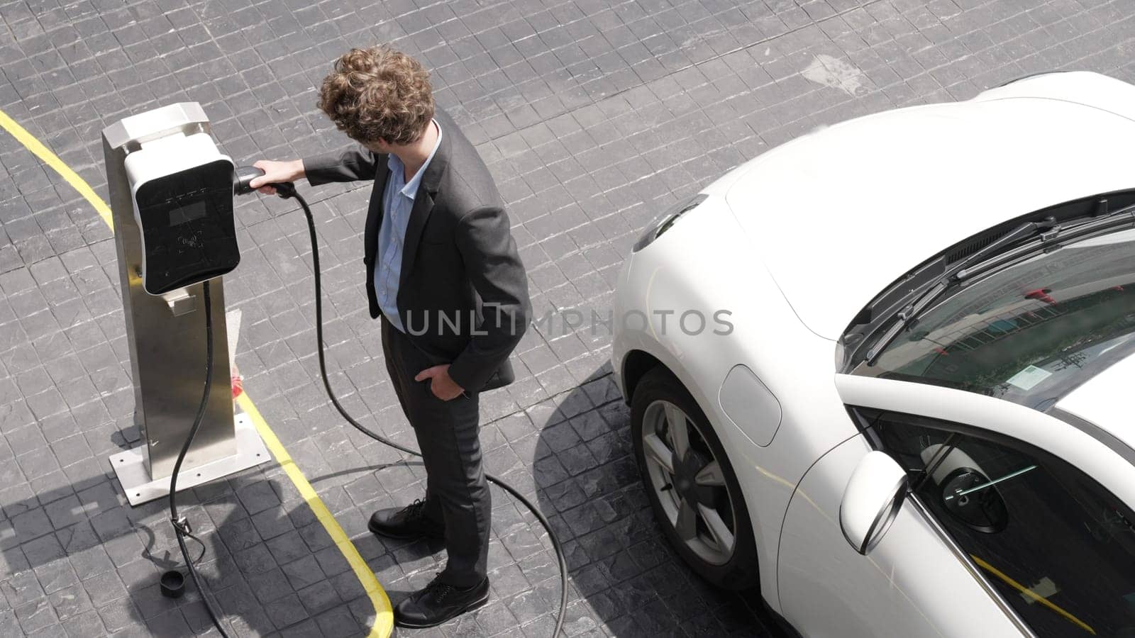 Progressive businessman install charger plug from charging station to his electric car before driving around city center. Eco friendly rechargeable car powered by sustainable and clean energy.