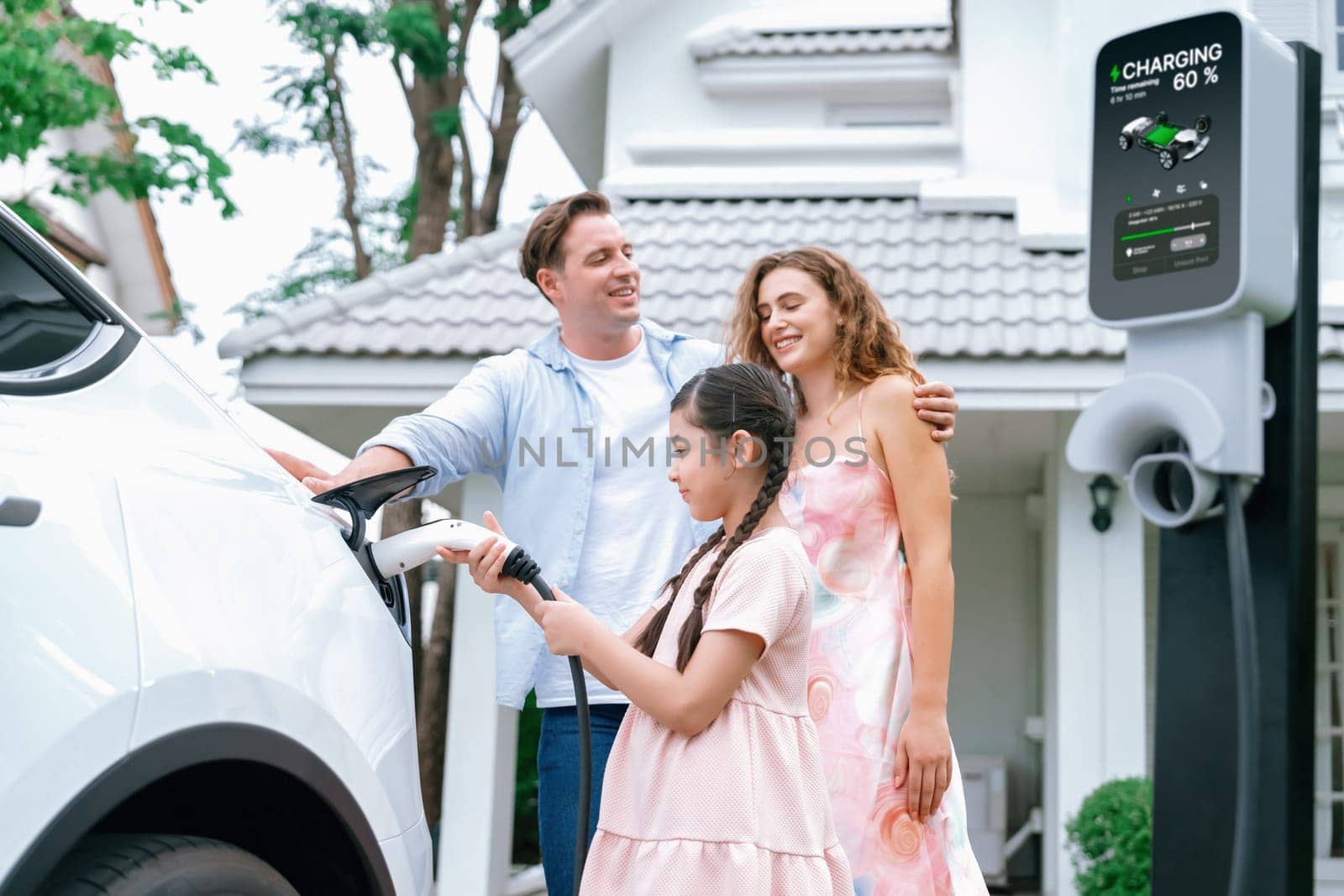 Happy little young girl learn about eco-friendly and energy sustainability as she help her family recharge electric vehicle from home EV charging station. EV car and modern family concept. Synchronos