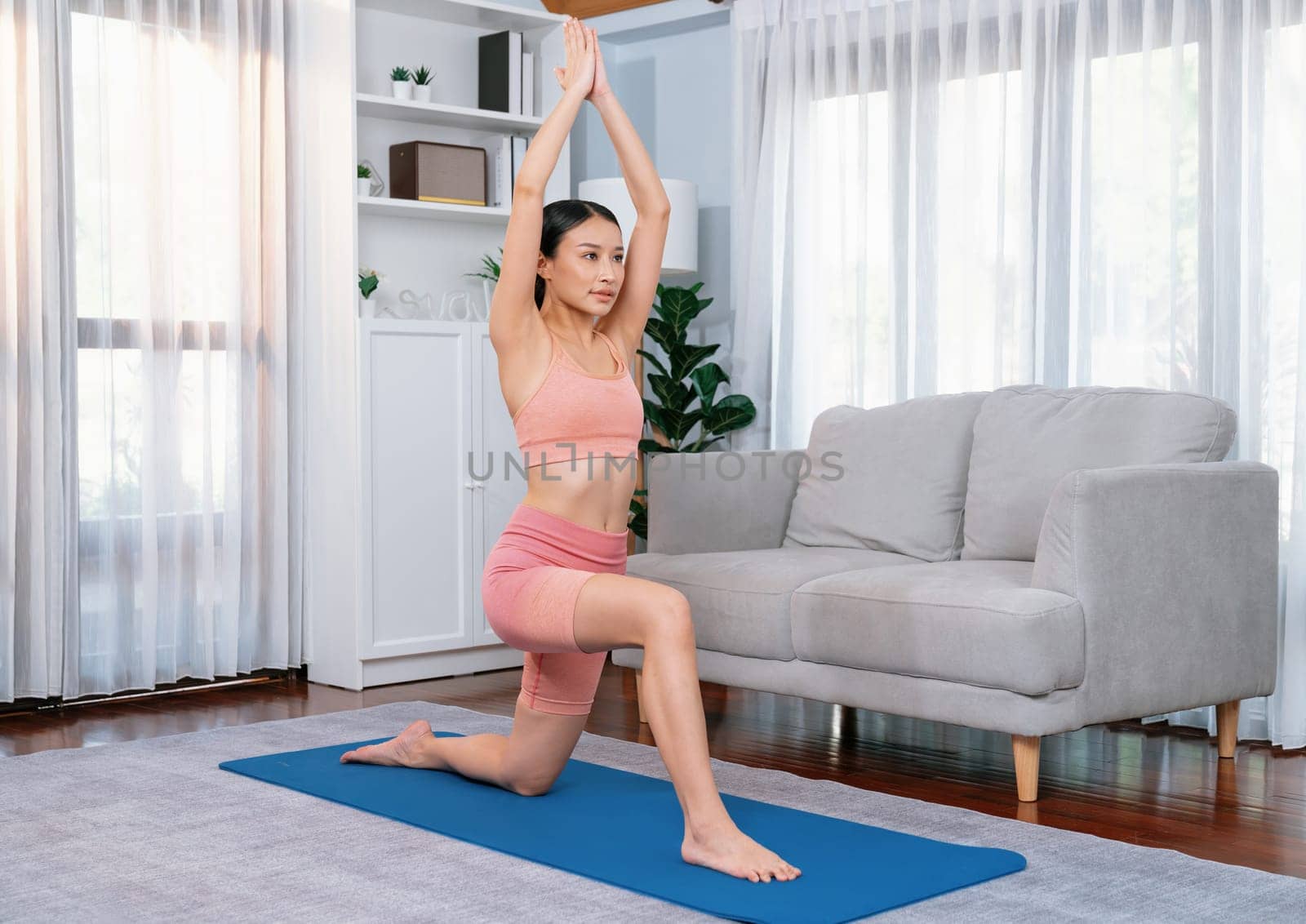 Asian woman in sportswear doing yoga exercise on fitness mat as her home workout training routine. Healthy body care and calm meditation in yoga lifestyle with comfortable and relaxation. Vigorous