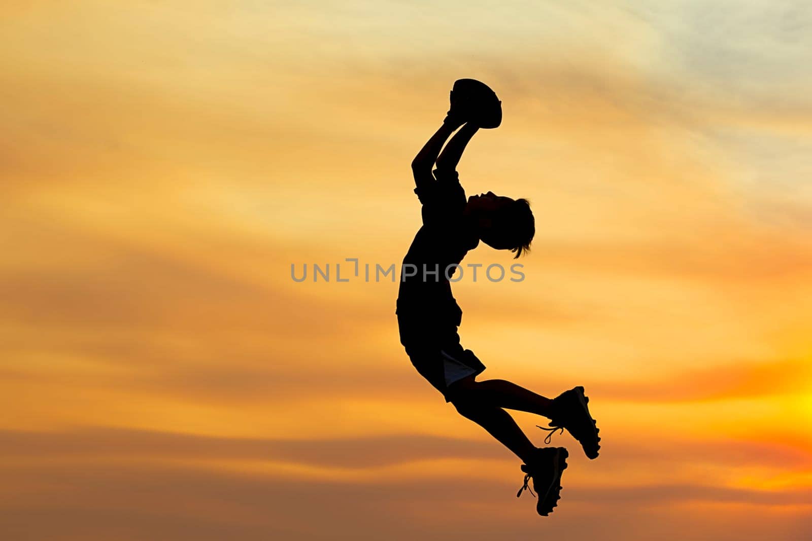 A young boy in silhouette is playing football at sunset against an orange sky.