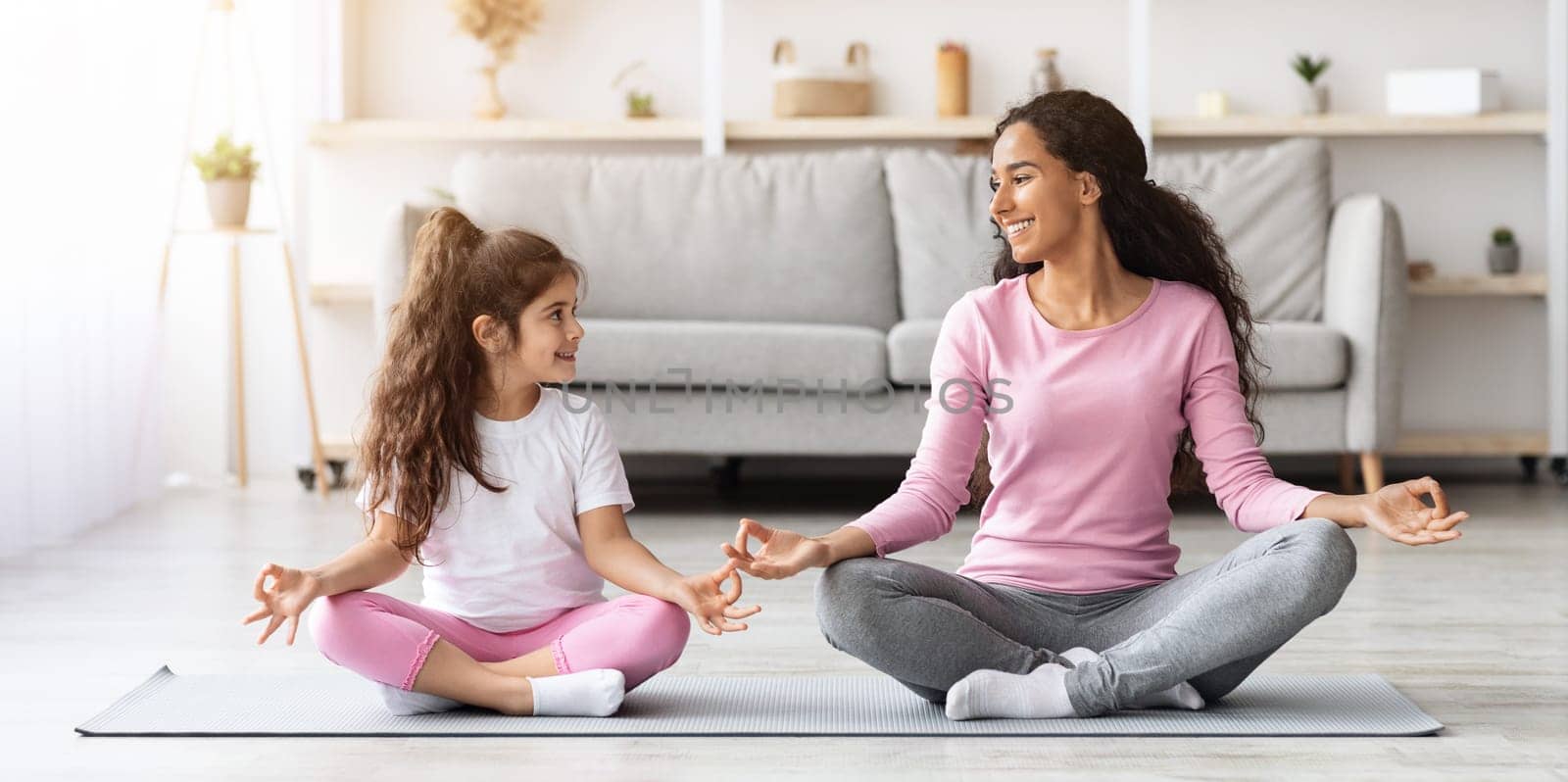 Cheerful young mother and little daughter in sportswear sitting on fitness mat in lotus position and smiling to each other, meditating together at home, copy space. Healthy family, sports lifestyle