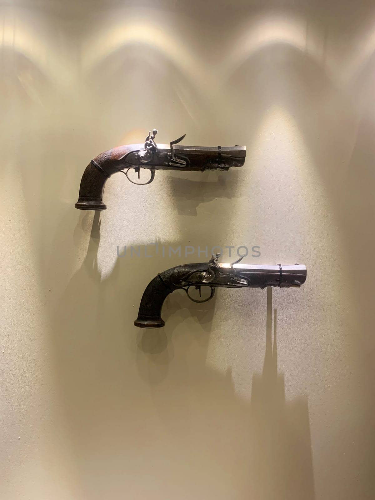 Two antique pistols hang on the wall, a lamp shines on them from above, forming bizarre shadows under them. High quality photo