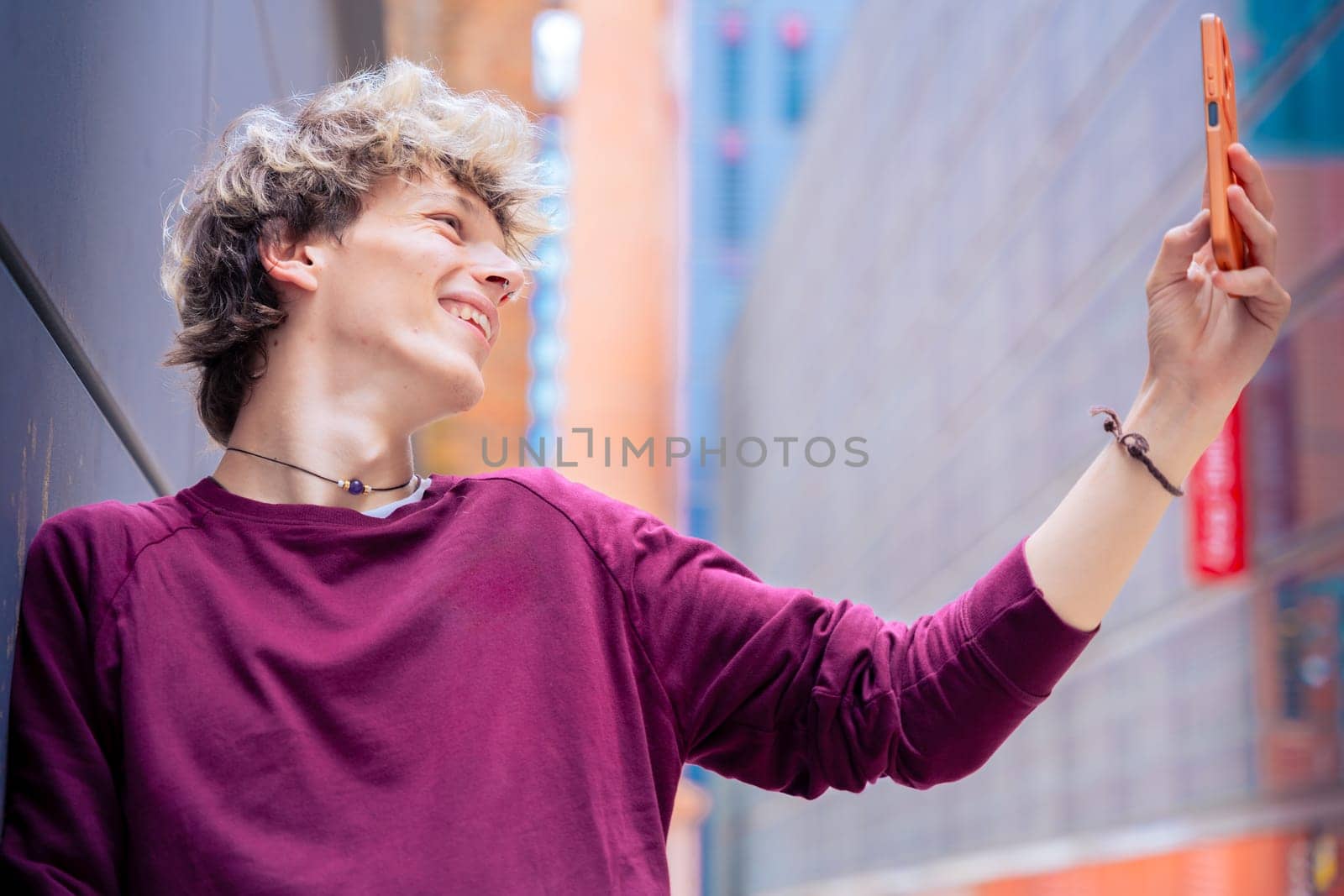 A cheerful young man with blond hair takes a selfie using his smartphone in a modern urban setting. Casual and vibrant scene highlighting youth and digital connectivity.