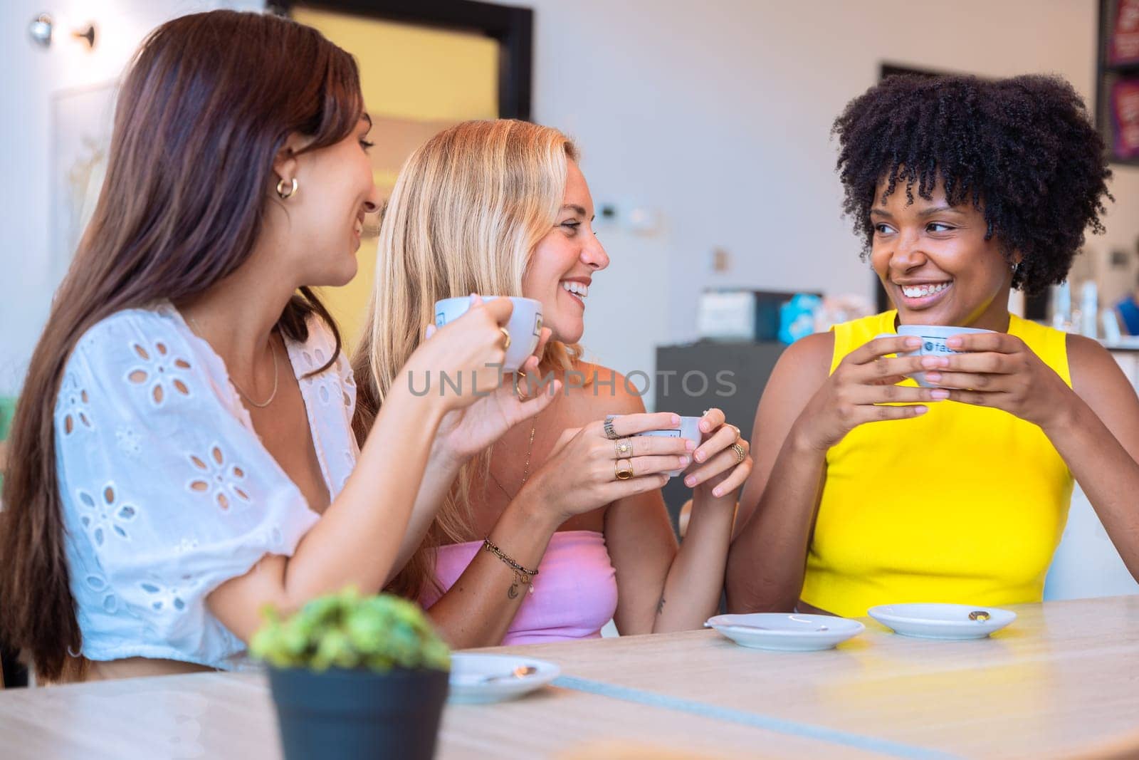 A group of three diverse women sitting at a table, holding coffee cups and laughing together in a cozy cafe. Warm and cheerful atmosphere, symbolizing friendship and connection.