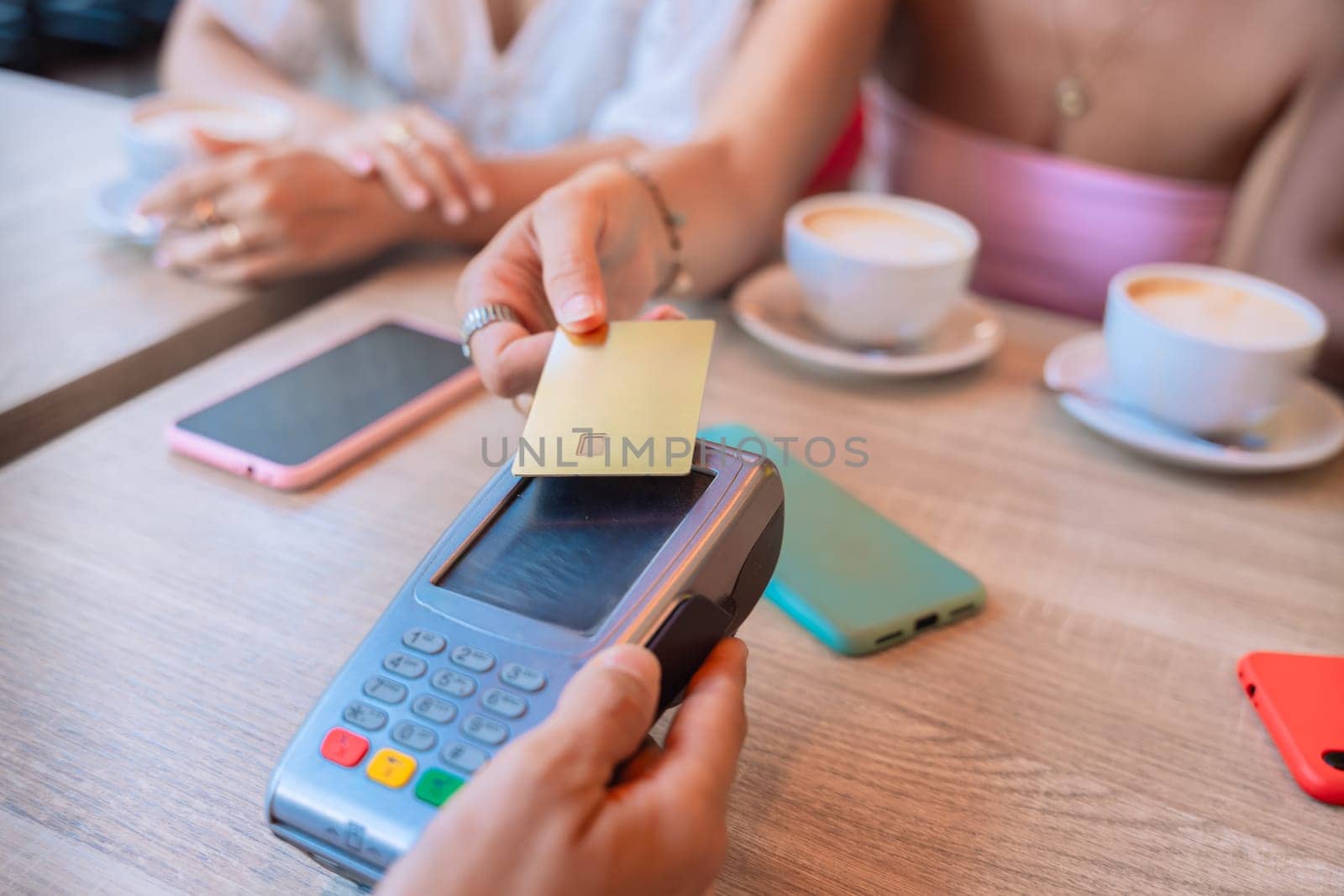 Close-up of a woman using a credit card for a contactless payment at a cafe. Coffee cups, smartphones, and a relaxed atmosphere in the background, symbolizing modern convenience.