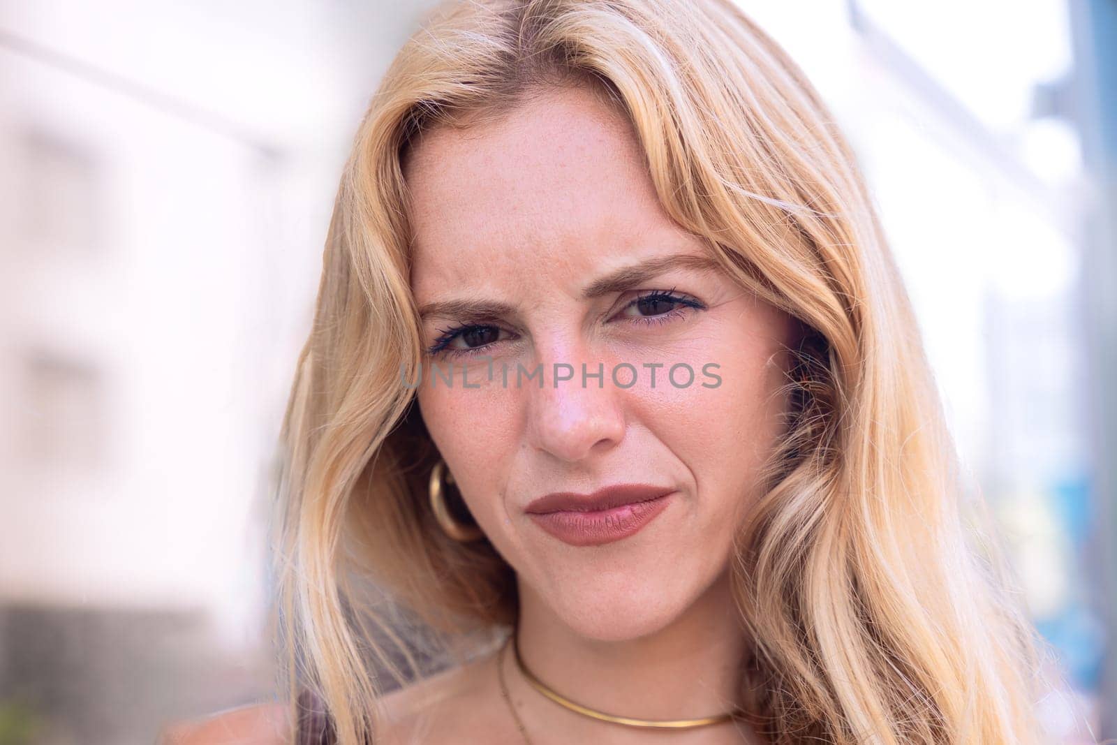 Portrait of a blonde woman with a serious, thoughtful expression. Focus on her facial features and natural beauty, captured outdoors with soft lighting and an urban background.