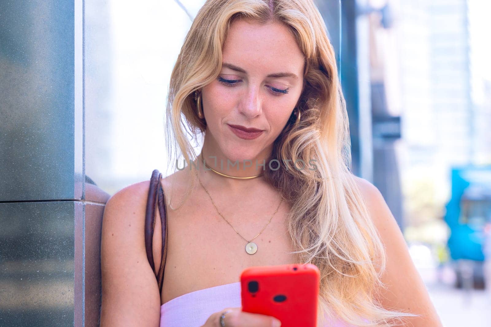 A blonde woman standing outdoors, focused on her smartphone. Captured in a modern urban environment, highlighting casual elegance, digital connectivity, and a relaxed lifestyle.