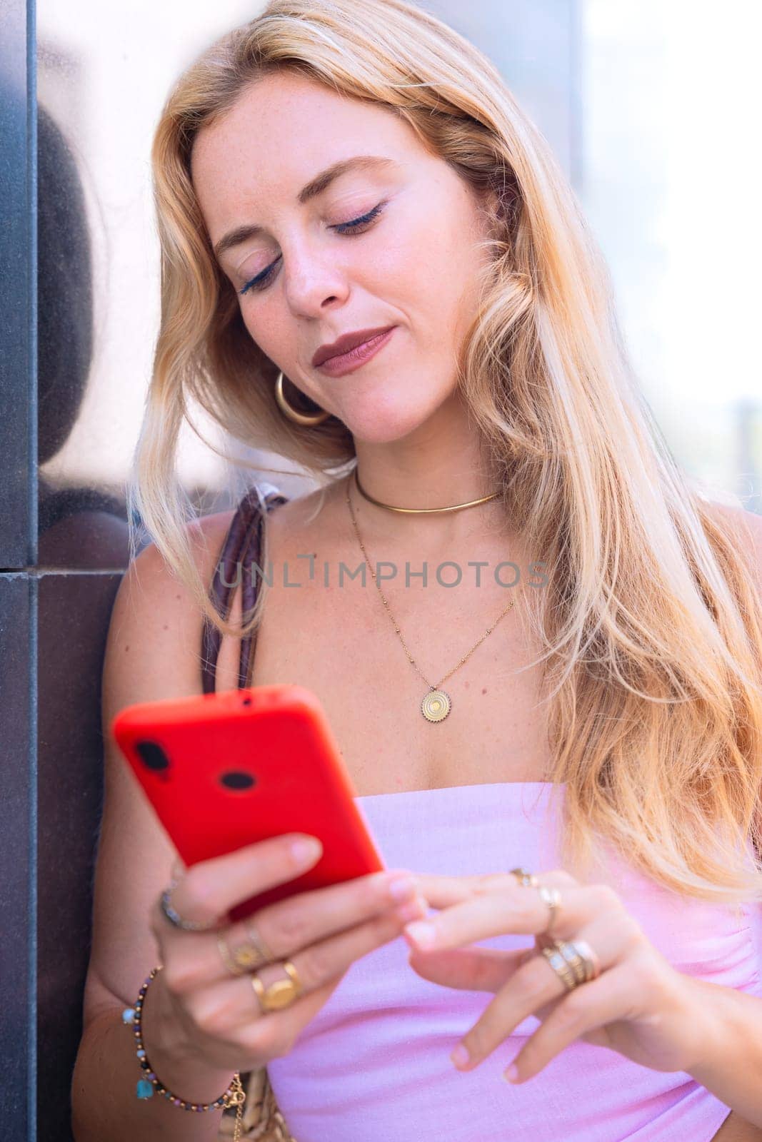 A blonde woman standing outdoors, focused on her smartphone. Captured in a modern urban environment, highlighting casual elegance, digital connectivity, and a relaxed lifestyle.