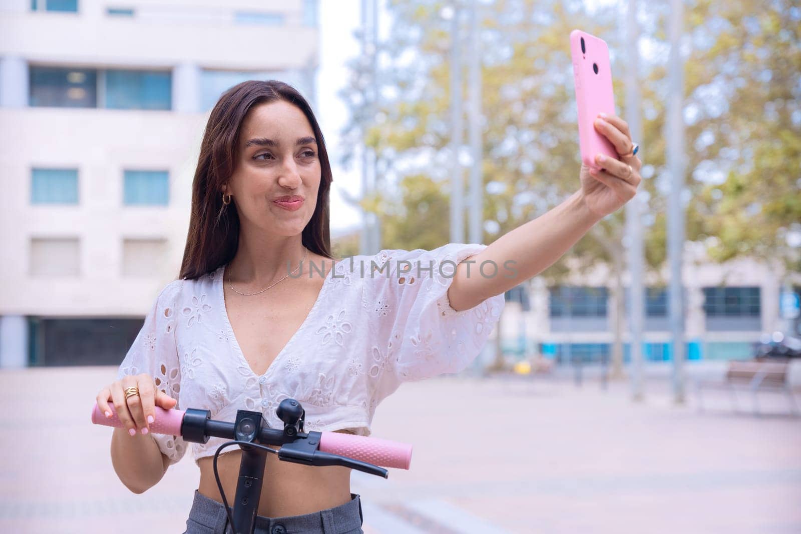 A stylish young woman with a cheerful expression takes a selfie outdoors while holding an electric scooter. Urban setting, showcasing modern mobility and digital connectivity.