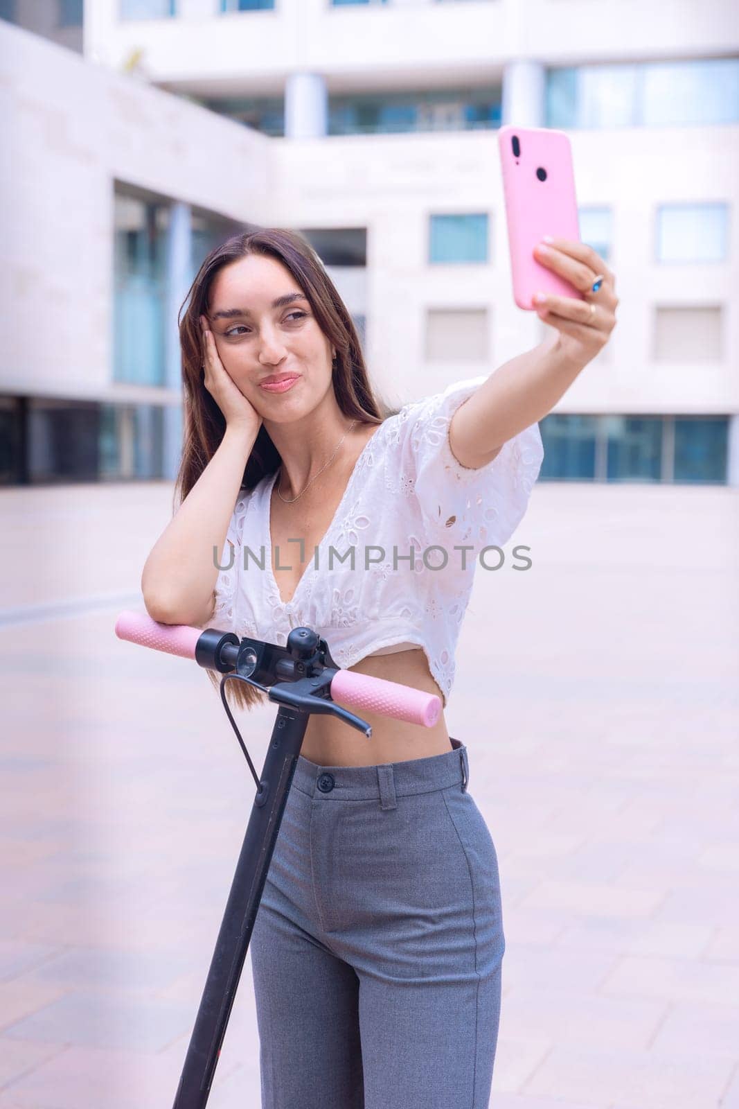A stylish young woman with a cheerful expression takes a selfie outdoors while holding an electric scooter. Urban setting, showcasing modern mobility and digital connectivity.
