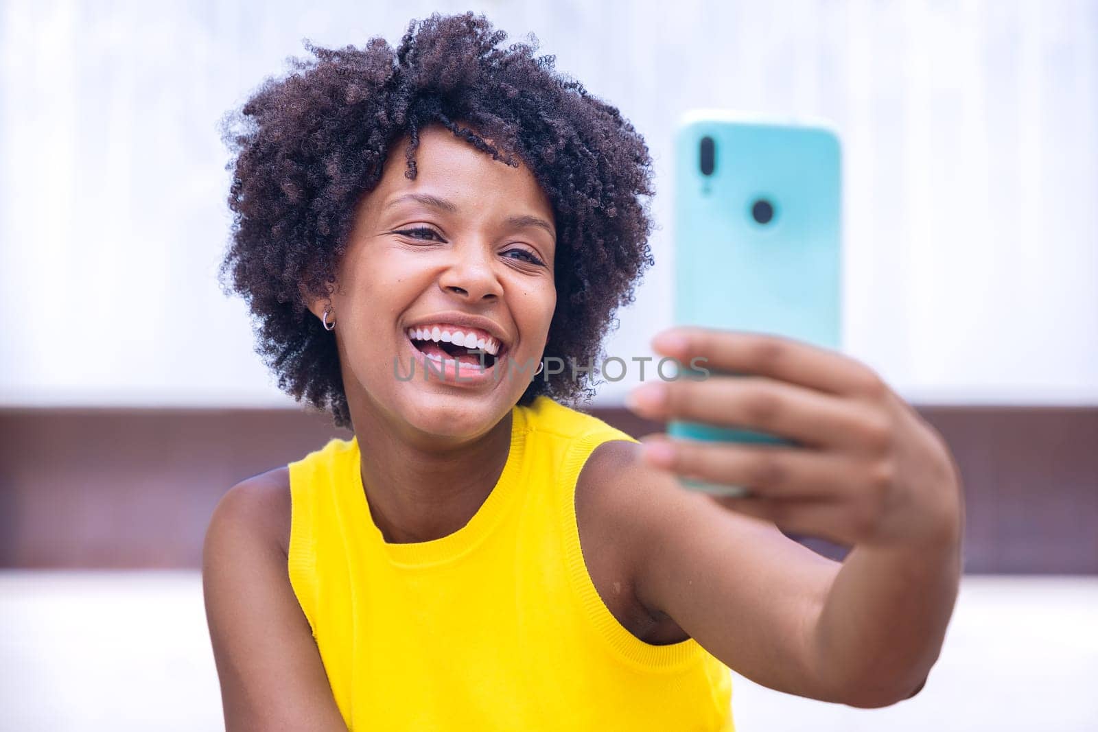 A joyful young woman with curly hair wearing a yellow top takes a selfie outdoors, smiling and gesturing peace. Vibrant scene showcasing happiness, individuality, and modern lifestyle.