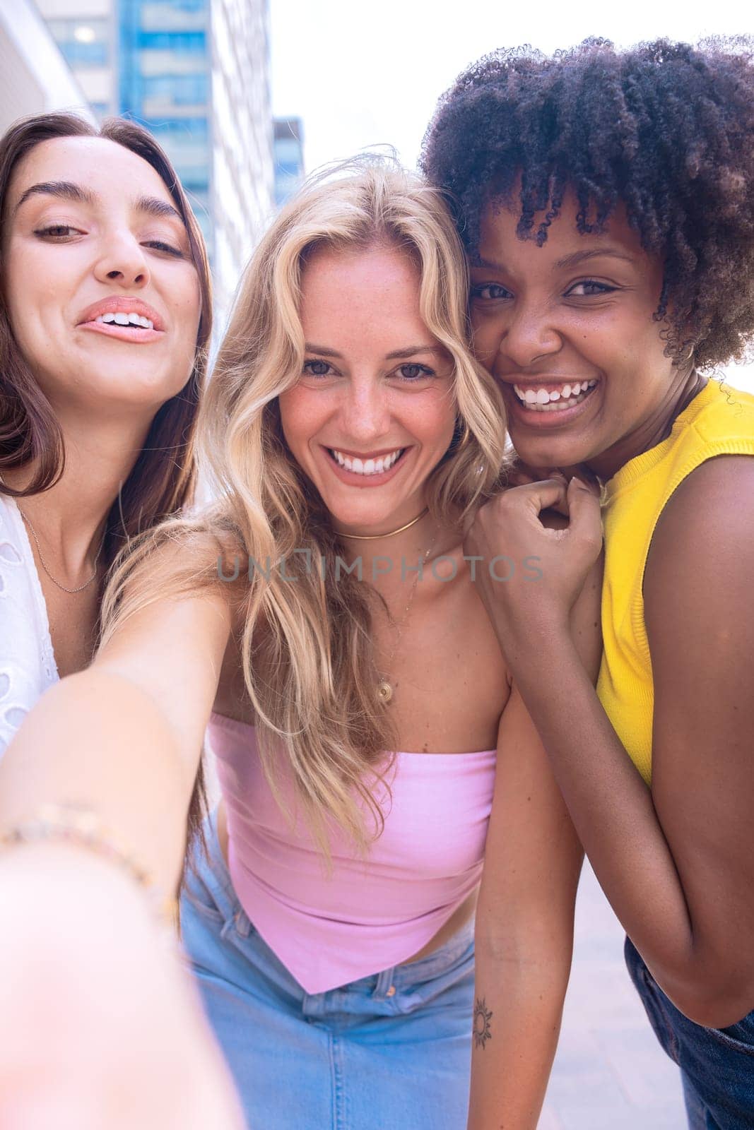 Three young women sitting outdoors, smiling, and taking a playful selfie together. Perfect for concepts like friendship, happiness, social media, and outdoor lifestyle.