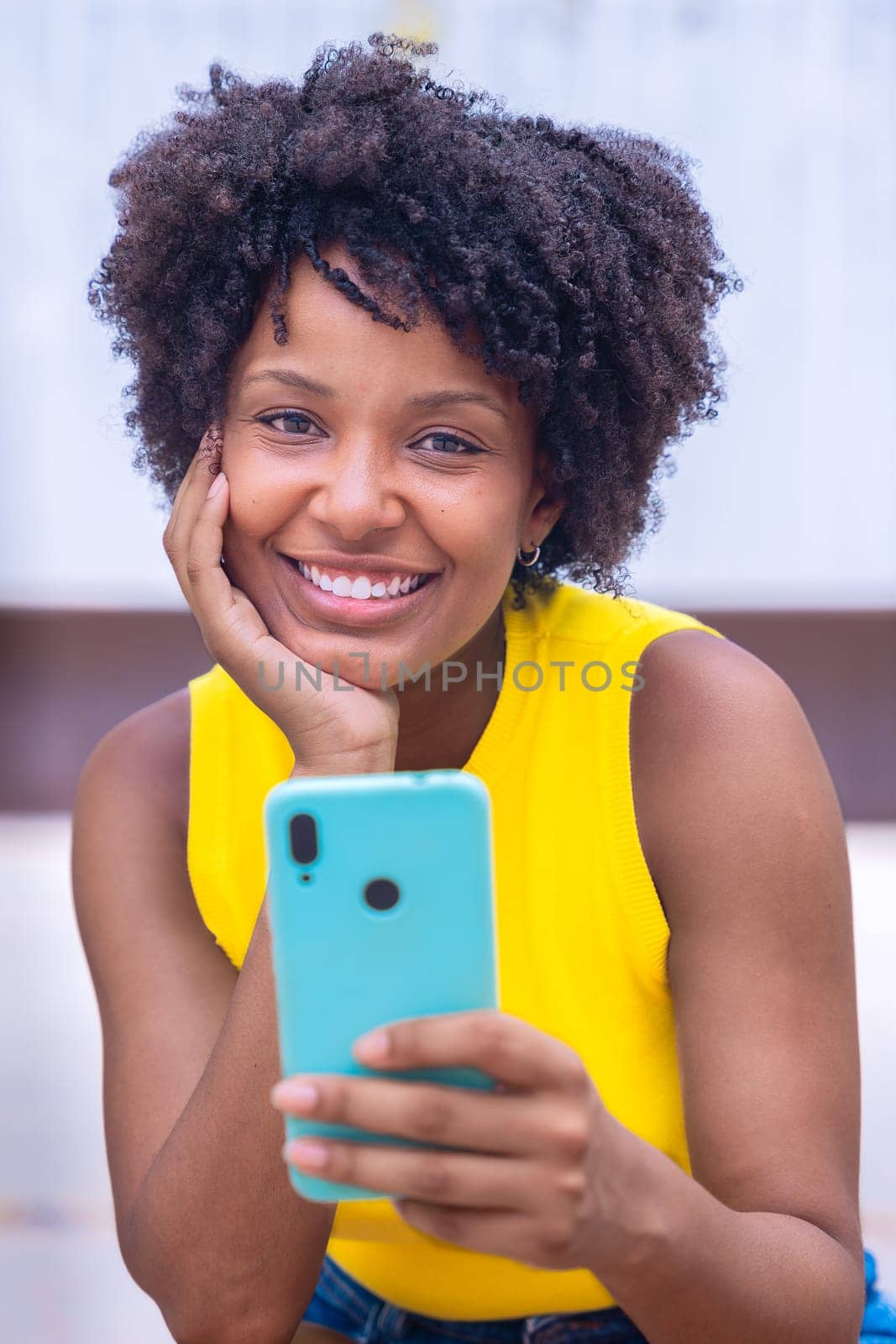 A young woman with natural curly hair wearing a yellow top rests her face on her hand while holding a smartphone. Calm and reflective moment captured in a bright outdoor setting.