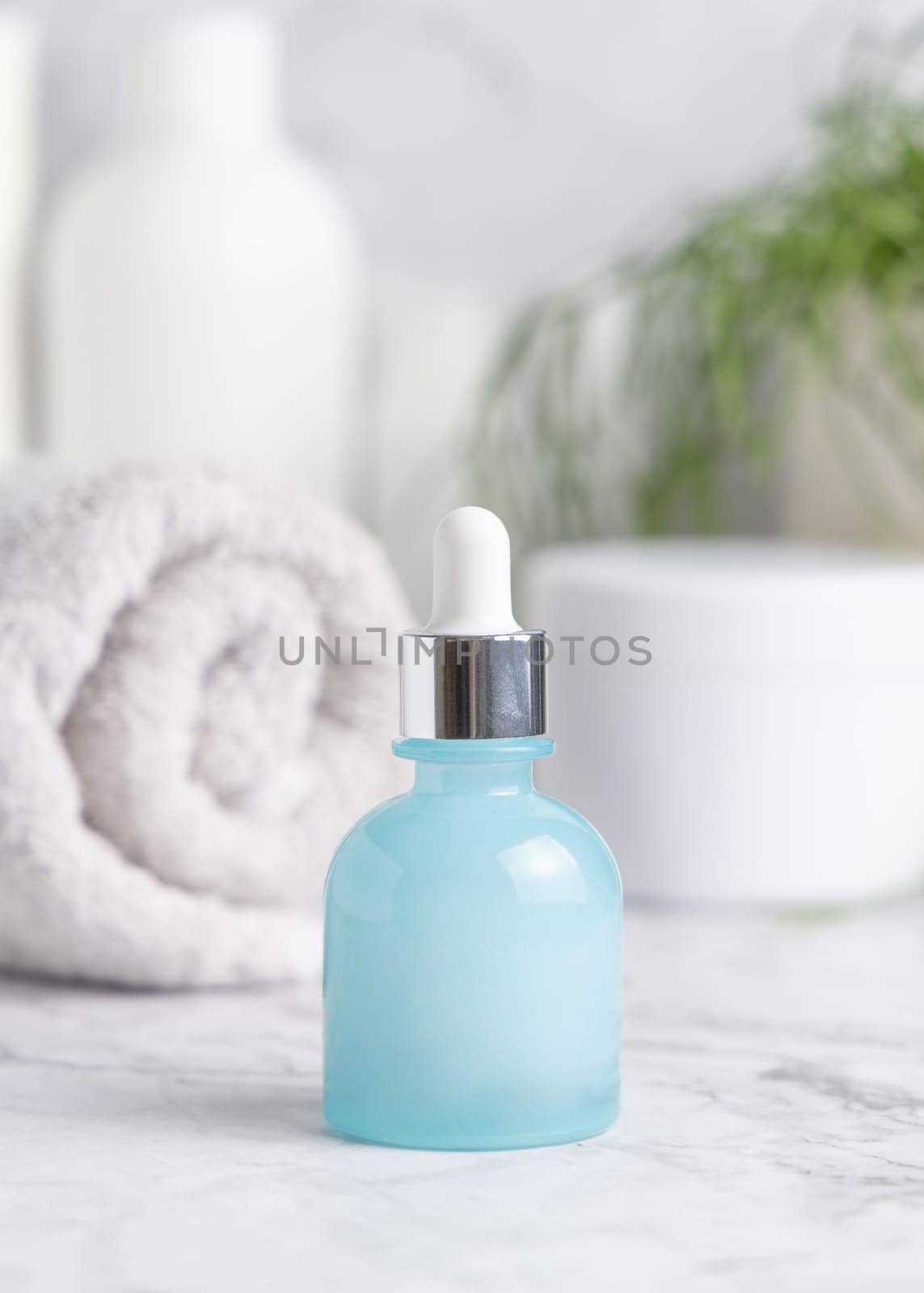 Light blue glass dropper bottle on white marble against green plant, towel and cosmetics in bathroom, close up, packaging mockup. Natural product for facecare, beauty routine