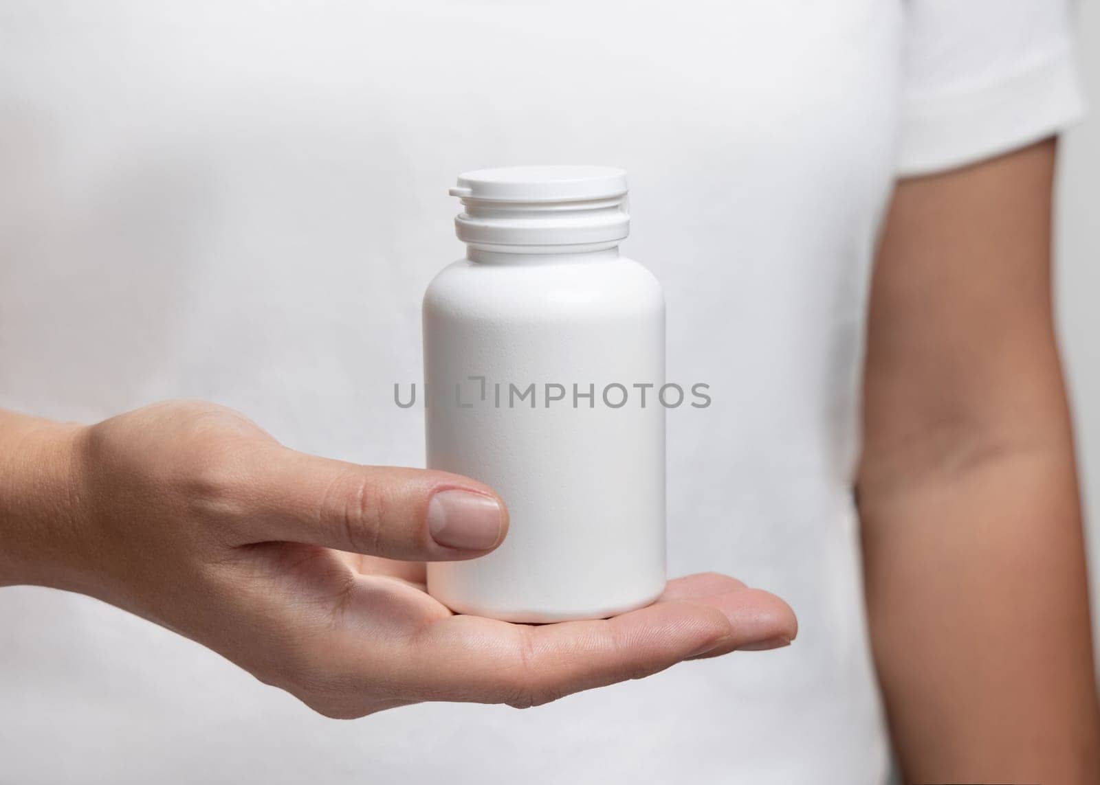 Closeup of woman in white t-shirt holding a white plastic medical pill bottle. Packaging mockup for herbal supplement. Female hands with natural manicure