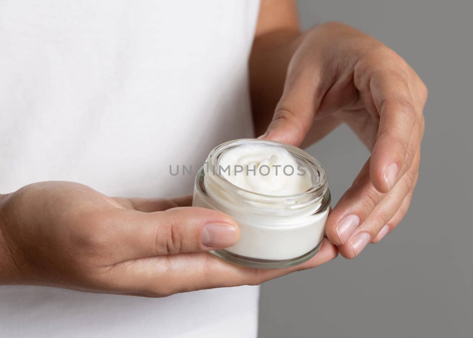 Female hand holding cream jar against white t-shirt close up. Natural product for facial care, everyday skincare routine. Opened cosmetic Container. Spa and Wellness concept