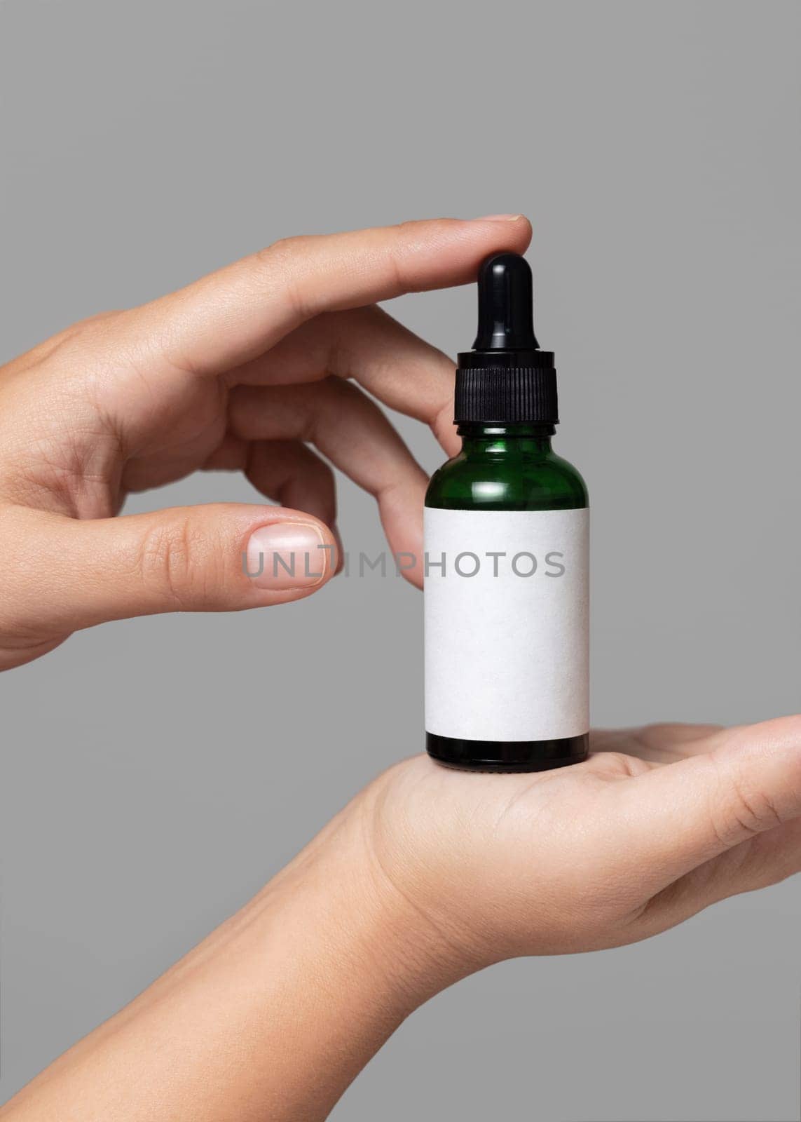 Female hands holding green dropper bottle with blank label against grey close up, cosmetic packaging mockup, copy space. Skincare beauty product - serum or essential oil