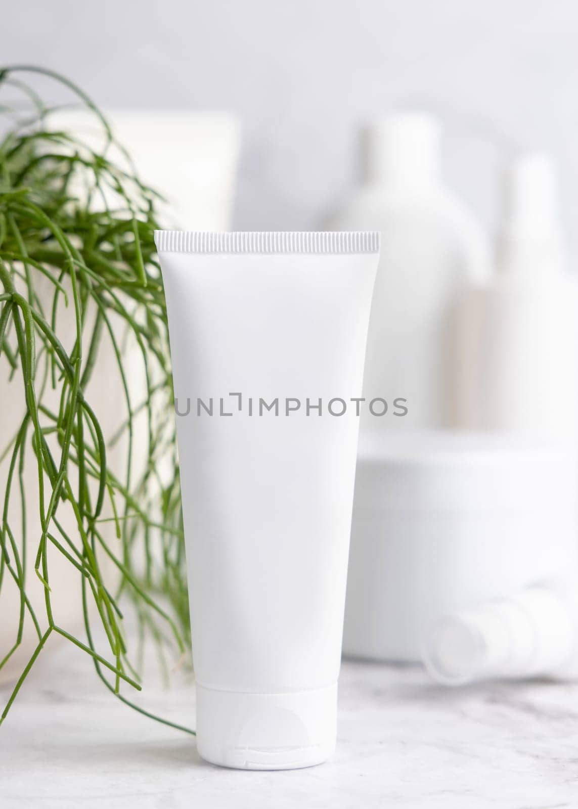 White cream squeeze tube on white marble table against green plant and cosmetic bottles in bathroom, close up, packaging mockup. Natural product for face care