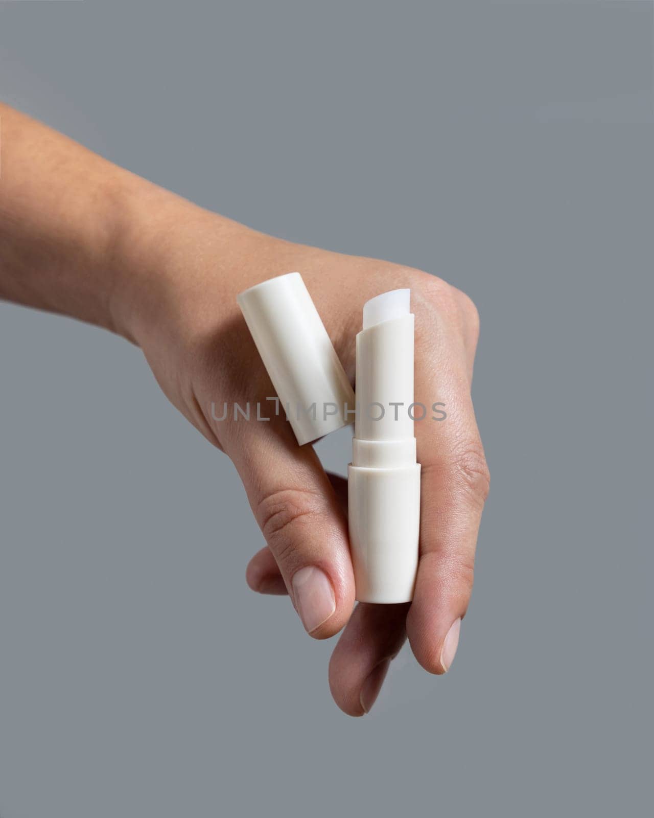 Female hand holding white opened lip balm stick with a lid against grey close up, cosmetic package mockup, copy space. Lip care beauty product with minimal composition