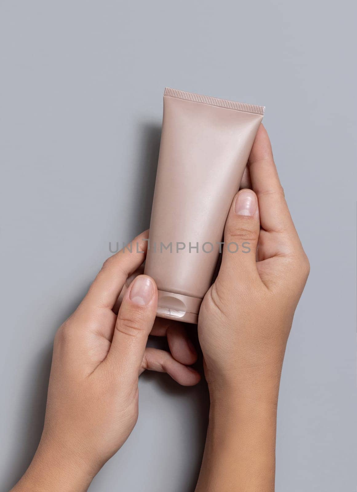 Female hands holding beige squeeze cream tube against grey close up, cosmetic package mockup, copy space. Skincare beauty product with minimal composition