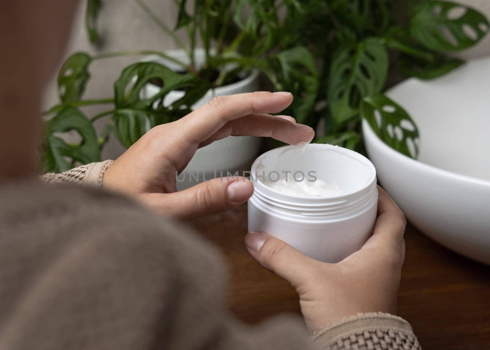 Close up of woman in brown bathrobe touching cream with finger and holding a cosmetic jar in other hand in bathroom against green plant and vessel basin. Shoulder view