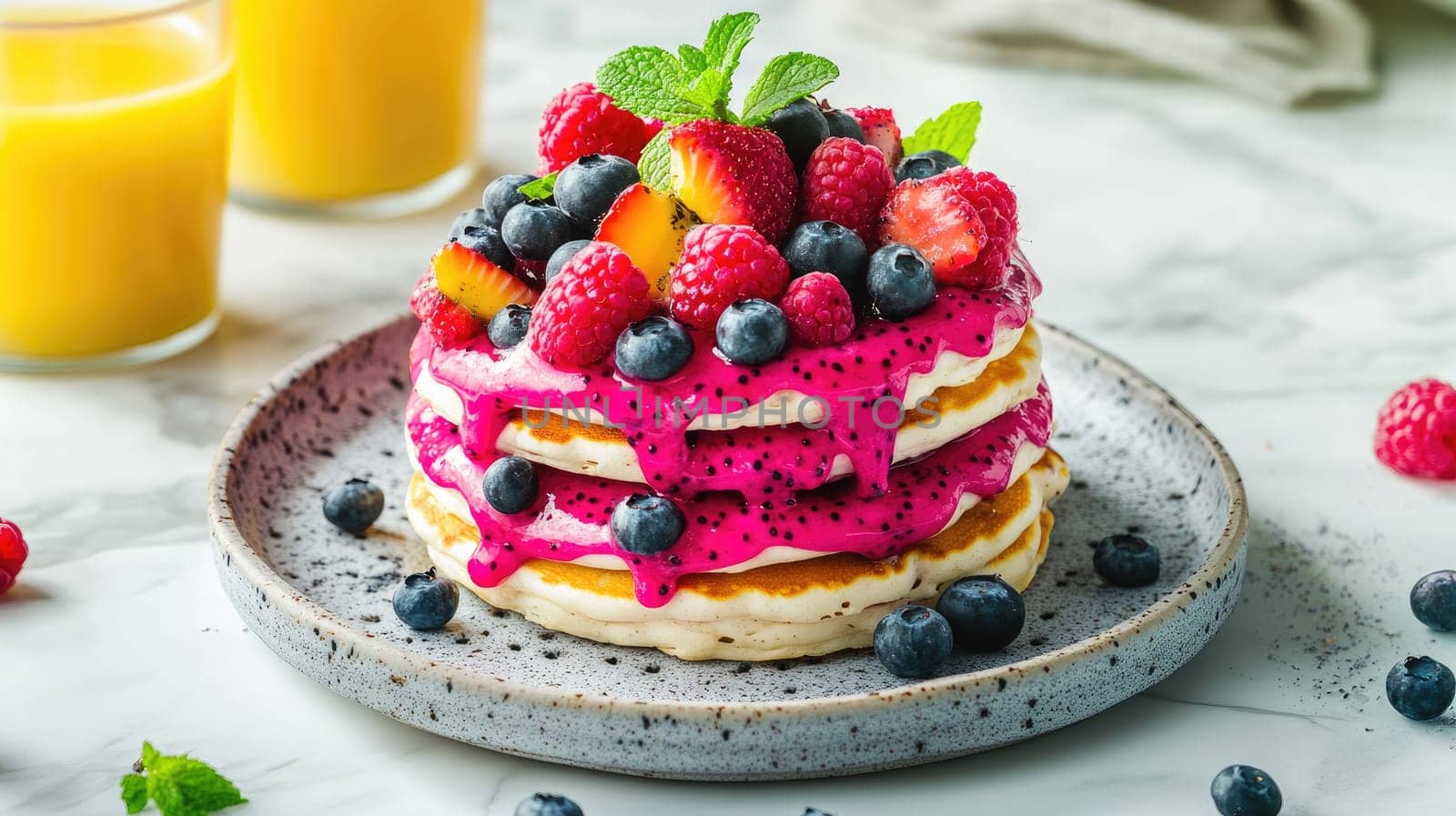 A delicious stack of fluffy pancakes layered with fresh berries and a bright pink fruity sauce garnished with mint. Two glasses of juice accompany the dessert.