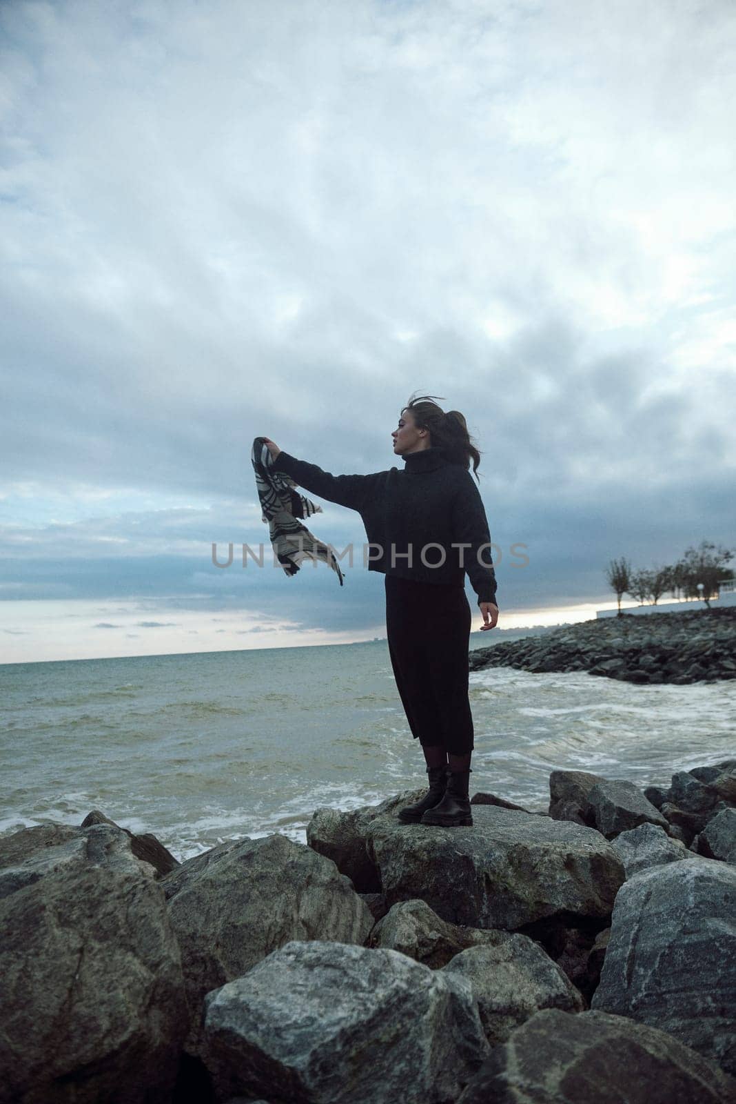 young girl on the seashore around the rocks. she is dressed in a beige trench coat, she is dreaming on the seashore among the flowers of the chrysanthemum variety. a chair decorated with blue fabric