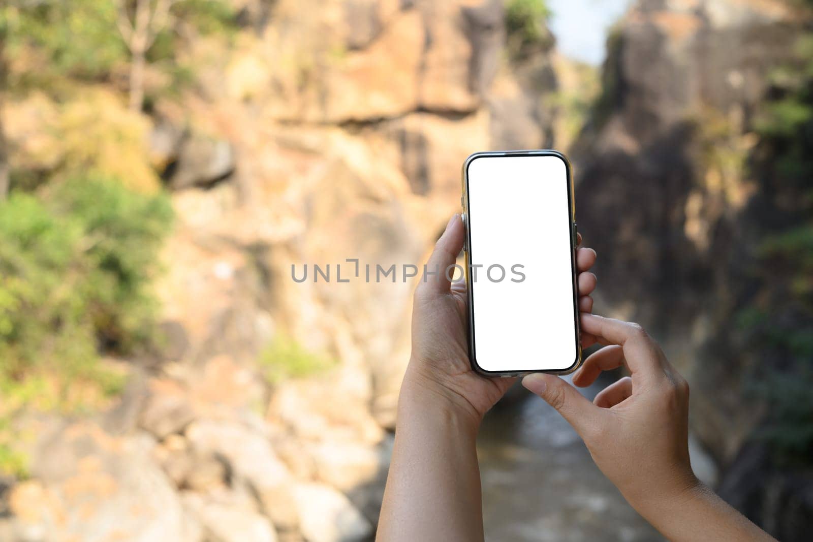 Close up of hands holding smartphone with blank screen against scenic outdoor backdrop of rocky terrain and greenery.