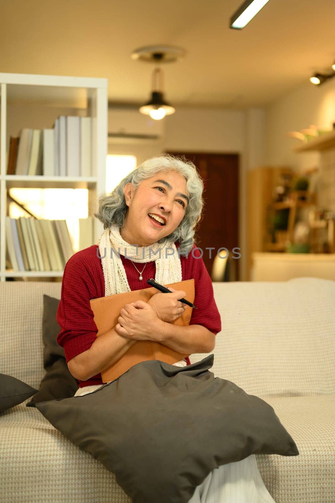 Calm senior woman in a red sweater sitting on couch and writing in journal.