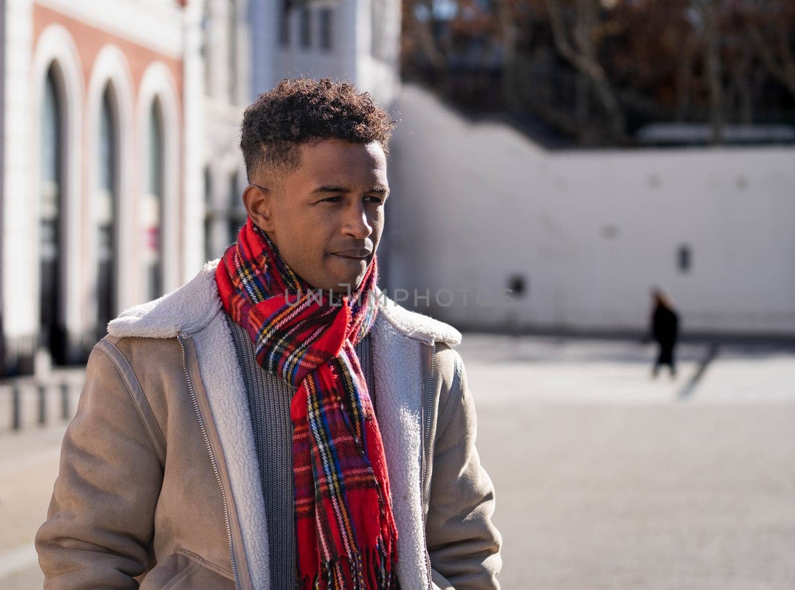Stylish black man wearing a red scarf and a beige jacket is walking in the city during a sunny winter day