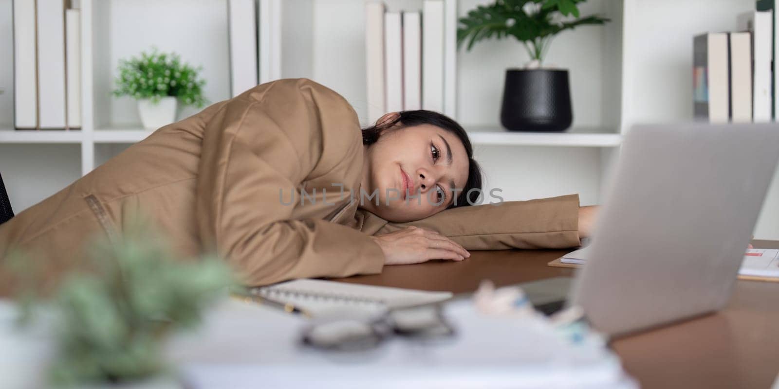 A businesswoman appears stressed and frustrated as she leans on her desk, surrounded by paperwork and her laptop, capturing a moment of challenge in her workday.