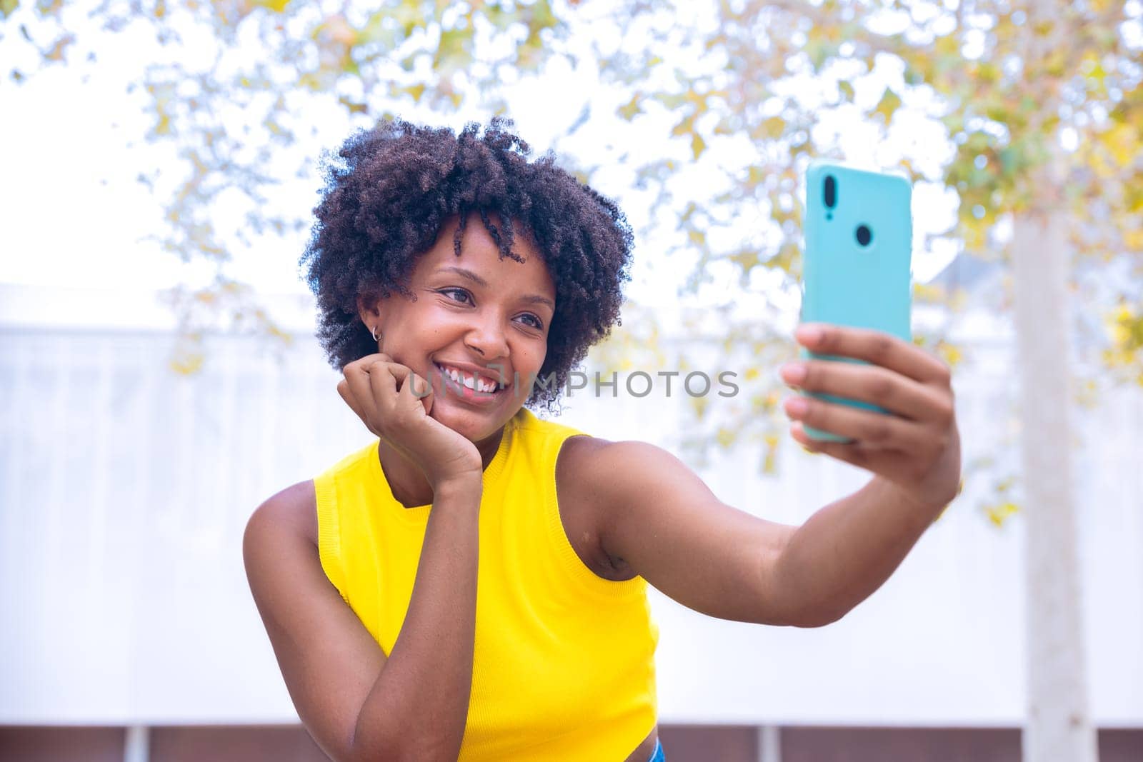 A joyful young woman with curly hair wearing a yellow top takes a selfie outdoors, smiling and gesturing peace. Vibrant scene showcasing happiness, individuality, and modern lifestyle.
