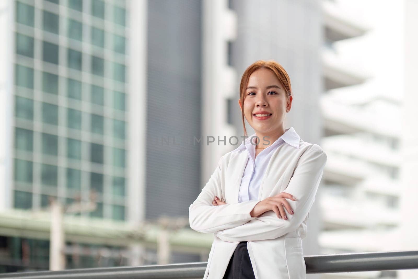 A self-assured businesswoman in a white blazer stands proudly with crossed arms, smiling against a backdrop of a sleek, contemporary office structure.