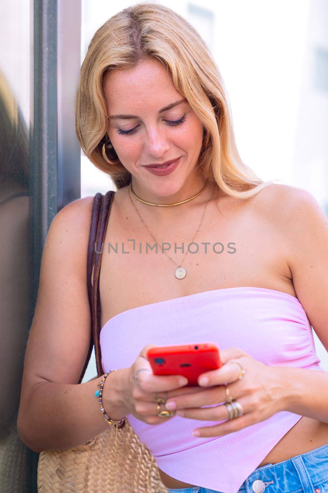 A blonde woman standing outdoors, focused on her smartphone. Captured in a modern urban environment, highlighting casual elegance, digital connectivity, and a relaxed lifestyle.