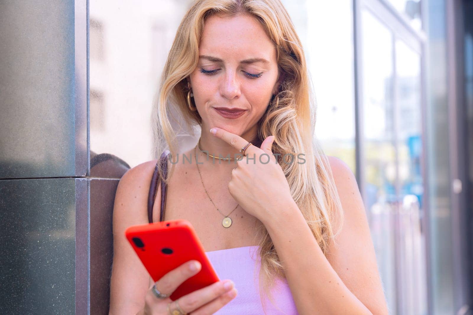 A blonde woman standing outdoors, focused on her smartphone. Captured in a modern urban environment, highlighting casual elegance, digital connectivity, and a relaxed lifestyle.