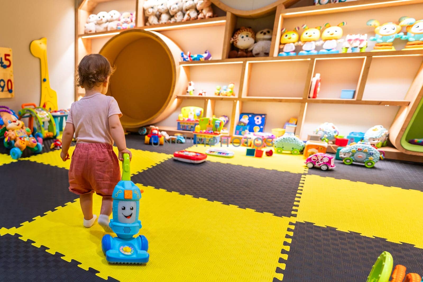 Toddler playing with a toy vacuum cleaner in a colorful playroom filled with educational toys and creative activities