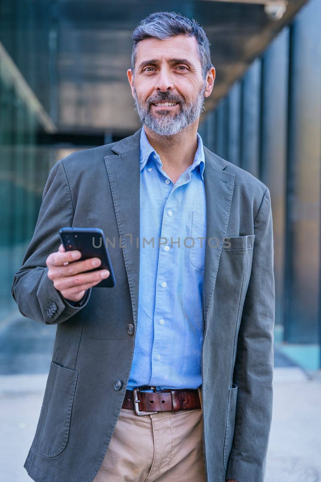 Smiling middle-aged businessman in a blue shirt holding and using a smartphone outdoors in a modern urban setting.