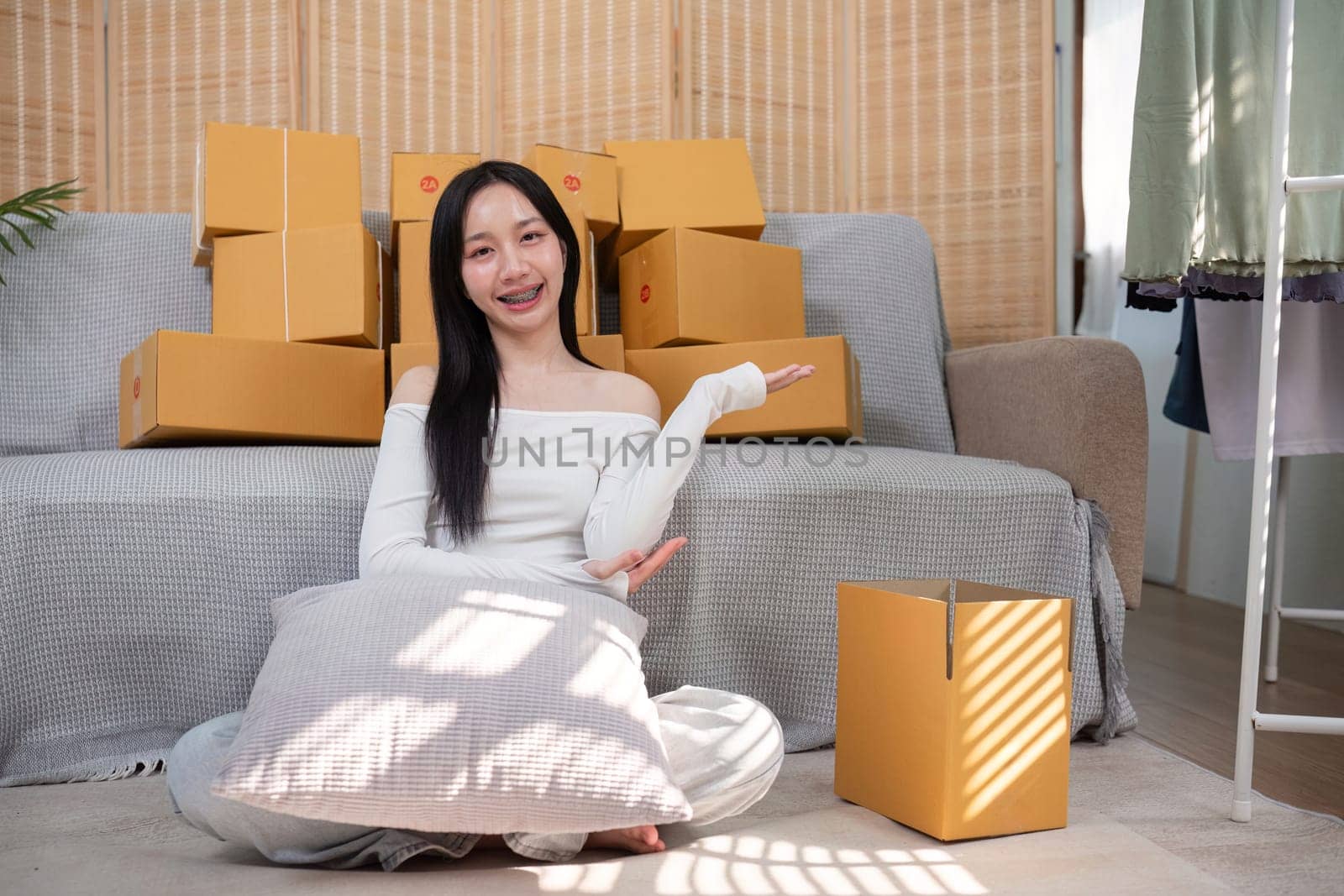 Delighted young woman with braces presenting her organized shipping area, surrounded by stacked packages and an empty box in her bright home workspace.