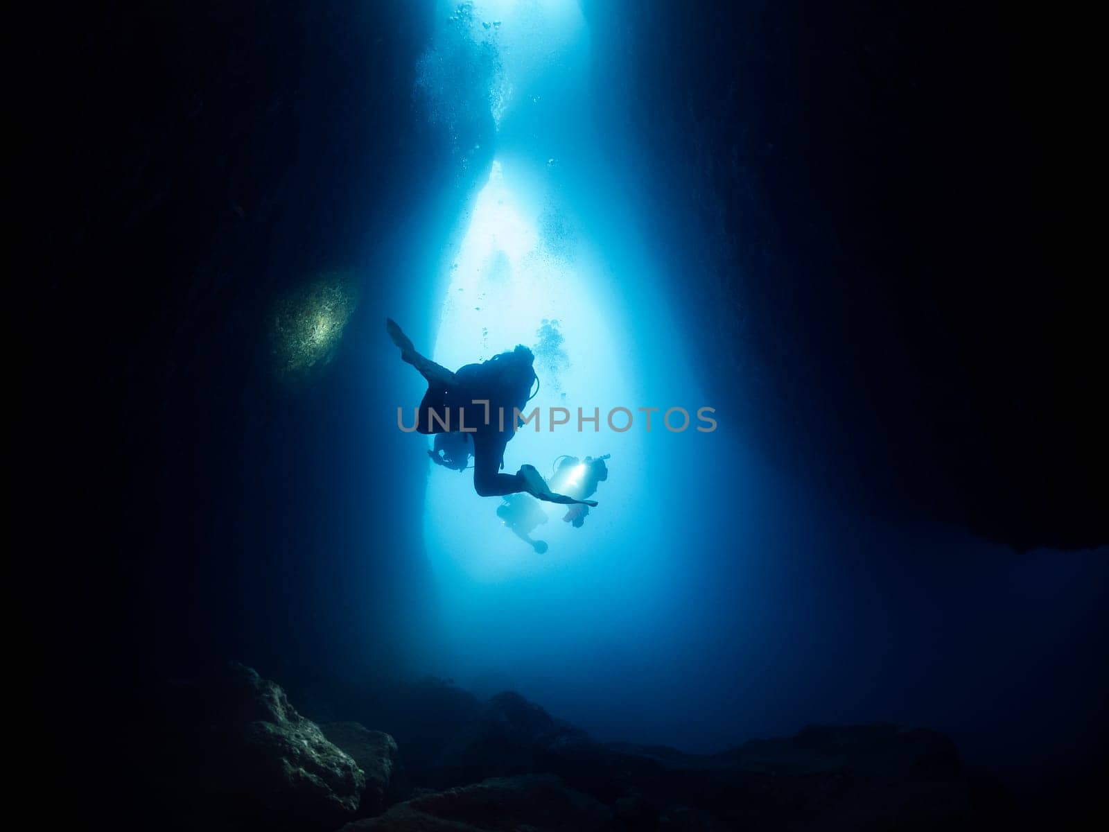 silhouette of unrecognizable scuba divers with flashlights swimming in narrow dark cave undersea in deep clear water, concept of active leisure and underwater adventure