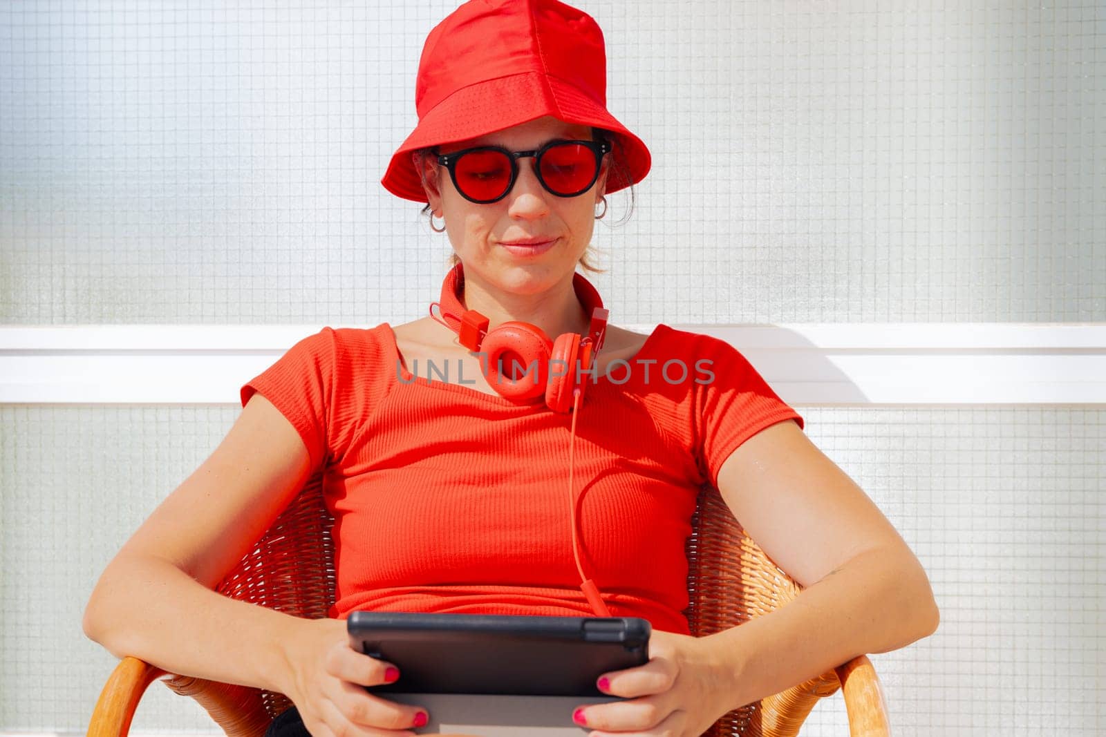 woman in a red hat, sunglasses, and headphones sits outdoors using a tablet. She enjoys a relaxing moment in the sun, dressed in a modern and vibrant outfit. The scene is bright and minimalistic.