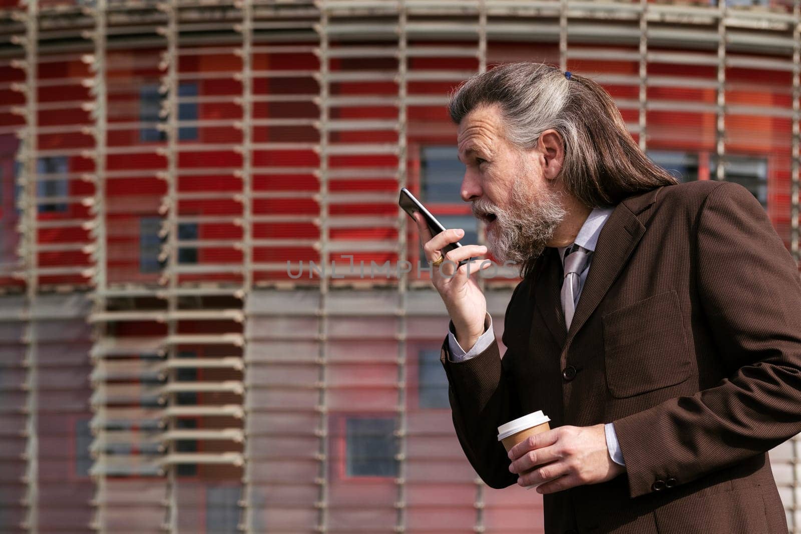 middle age male with beard in elegant suit with cup of takeaway coffee in hand talking on mobile phone against urban building, concept of business and mature man lifestyle, copyspace for text