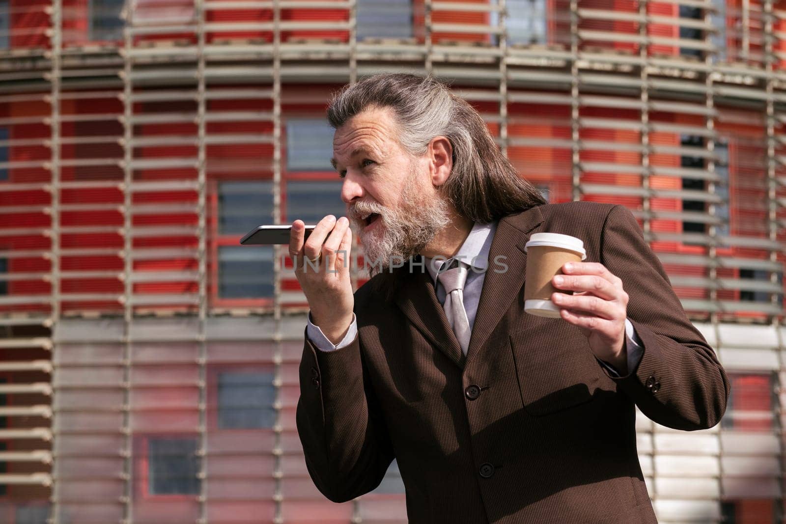 middle age male with beard in elegant suit with cup of takeaway coffee in hand recording voice message on mobile phone against urban building, concept of business and mature man lifestyle