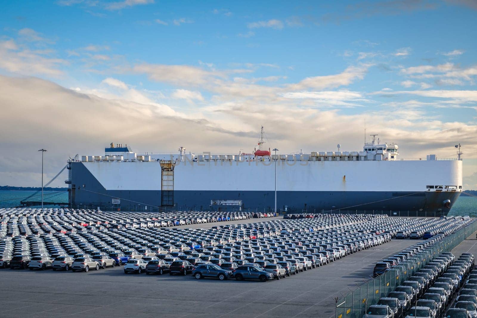 Large vehicle carrier ship known as RO-RO at port on sunny day, large parking area filled with cars. Blue sky and colorful clouds, vehicle transportation.